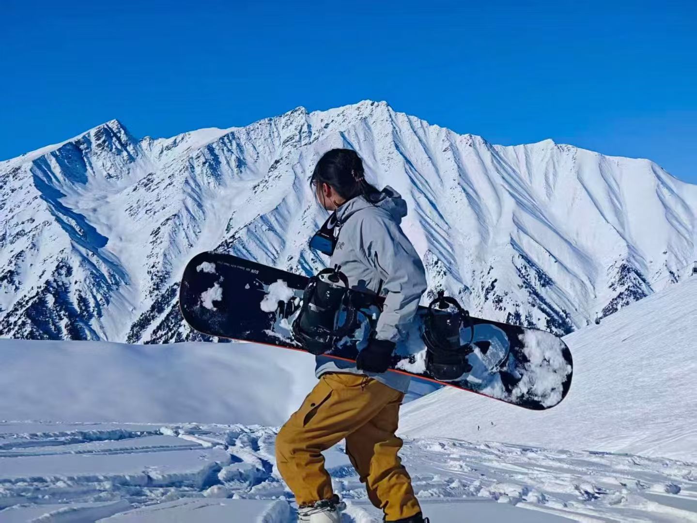 Shuyuan Hu carrying a snowboard in snowy mountains.